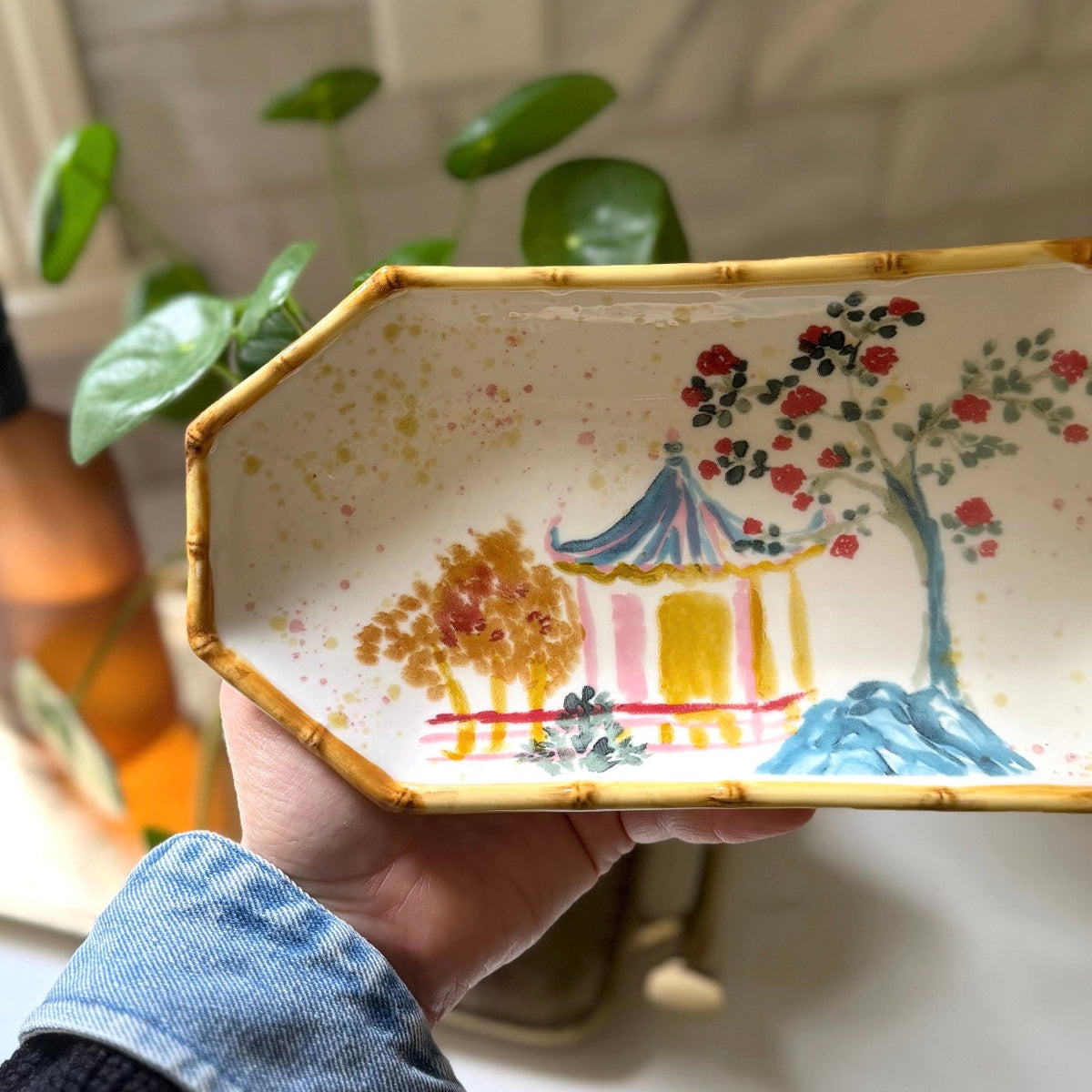 A hand holding a rectangular ceramic dish with an octagonal end, decorated with a hand-painted landscape scene featuring a pagoda, trees, and mountains. The dish has a gold rim and is held in front of a kitchen background with a tiled wall and a plant.